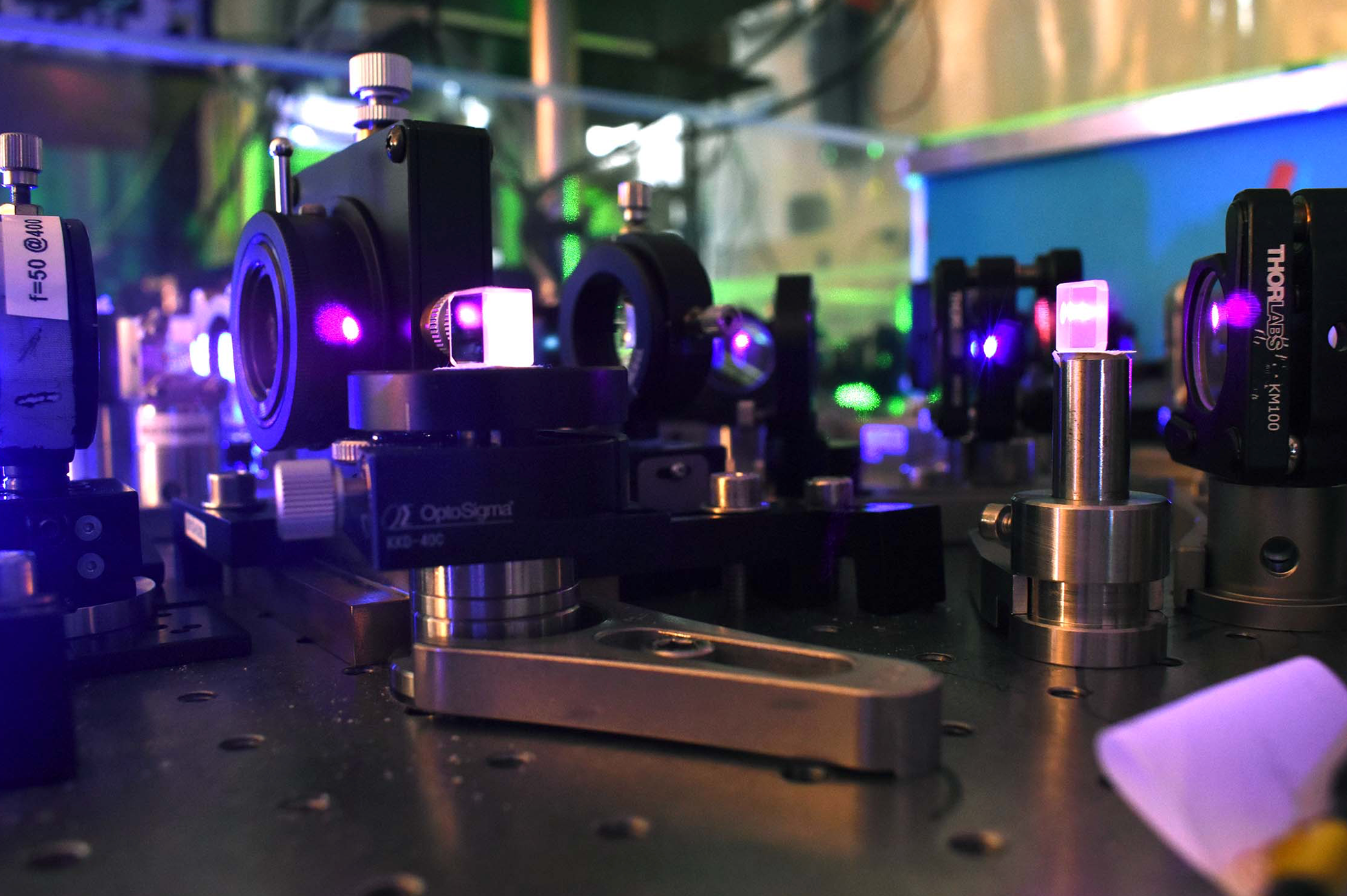 A close-up of the optical table side zoomed-in view of a quantum optics table, with several components locked on steel plate and emitting purple-like laser light.